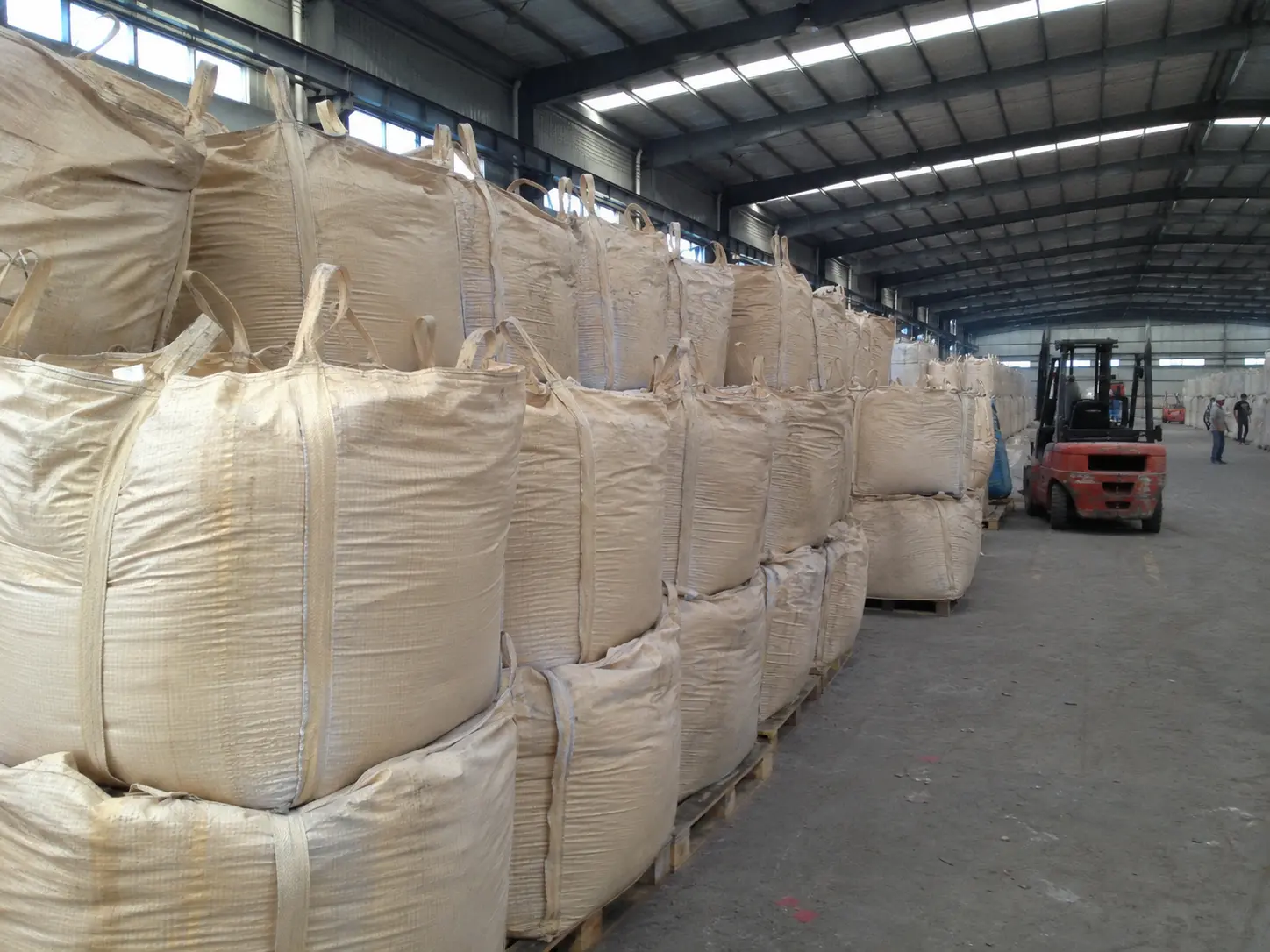 Rows of filled white and beige bulk bags stacked on pallets in a large warehouse with a forklift